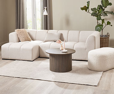 Moderni, muhkea beige sohva, tummanruskea lieriön mallinen sohvapöytä sekä teddykankainen ovaali rahi olohuoneessa