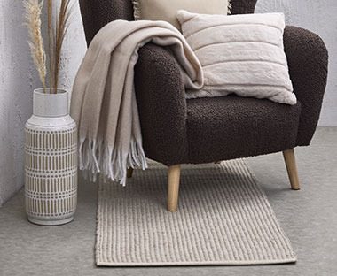 Harmaa nojatuoli jossa huopa, koristetyyny ja tyylikäs matto tuolin alla
