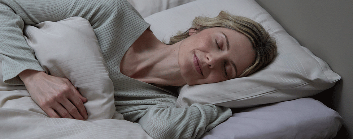 Nainen nukkuu kyljellään sängyssä valkoisella tyynyllä ja peitolla