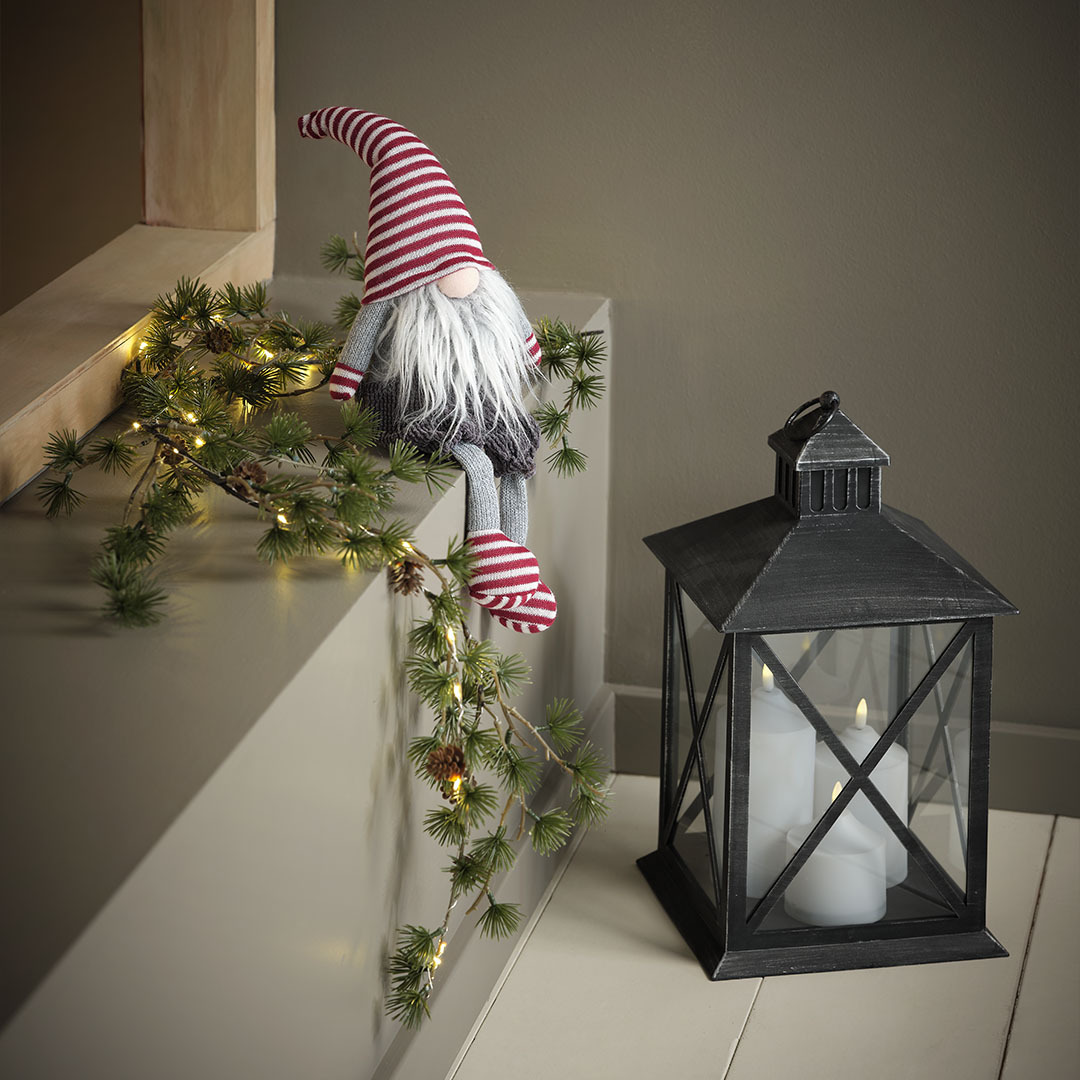 Joulutonttu, jolla on punavalkoiset lapaset, sukat ja tonttulakki, istumassa mustan lyhdyn vieressä