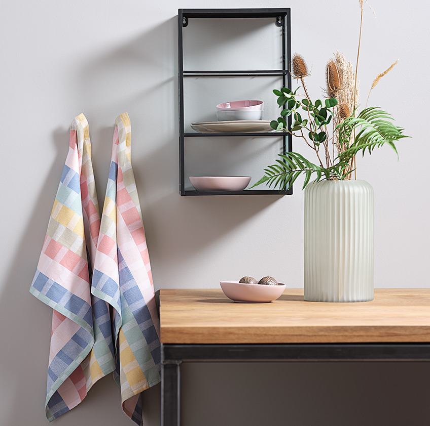 Cotton tea towels hanging next to a wall shelf and a vase on a table
