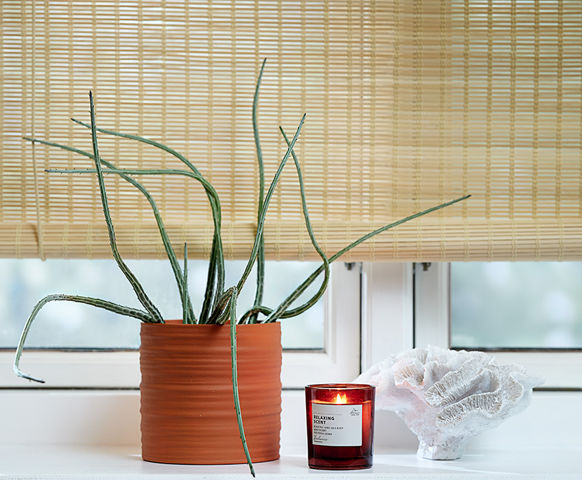 Bamboo roller blinds in a window with a plant pot, a scented candle and an ornament in the windowsill