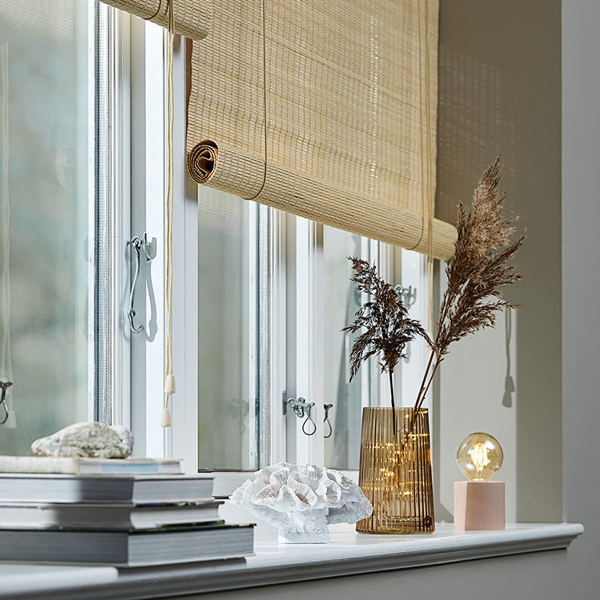 Bamboo roller blinds in a window with plant pot, ornament, vase, scented candle and battery lamp in the windowsill